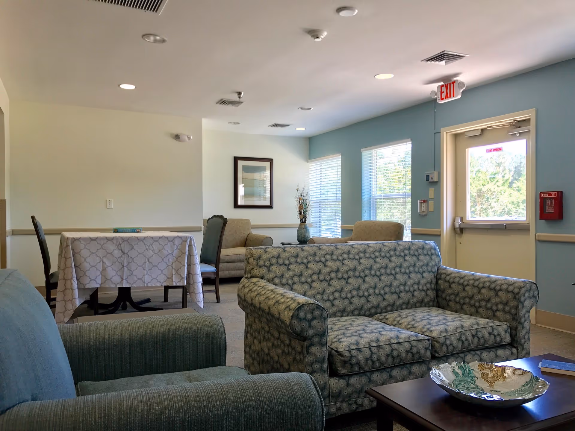 Sunlit common area with patterned sofas and armchairs, a table with a tablecloth, windows, and an exit door.