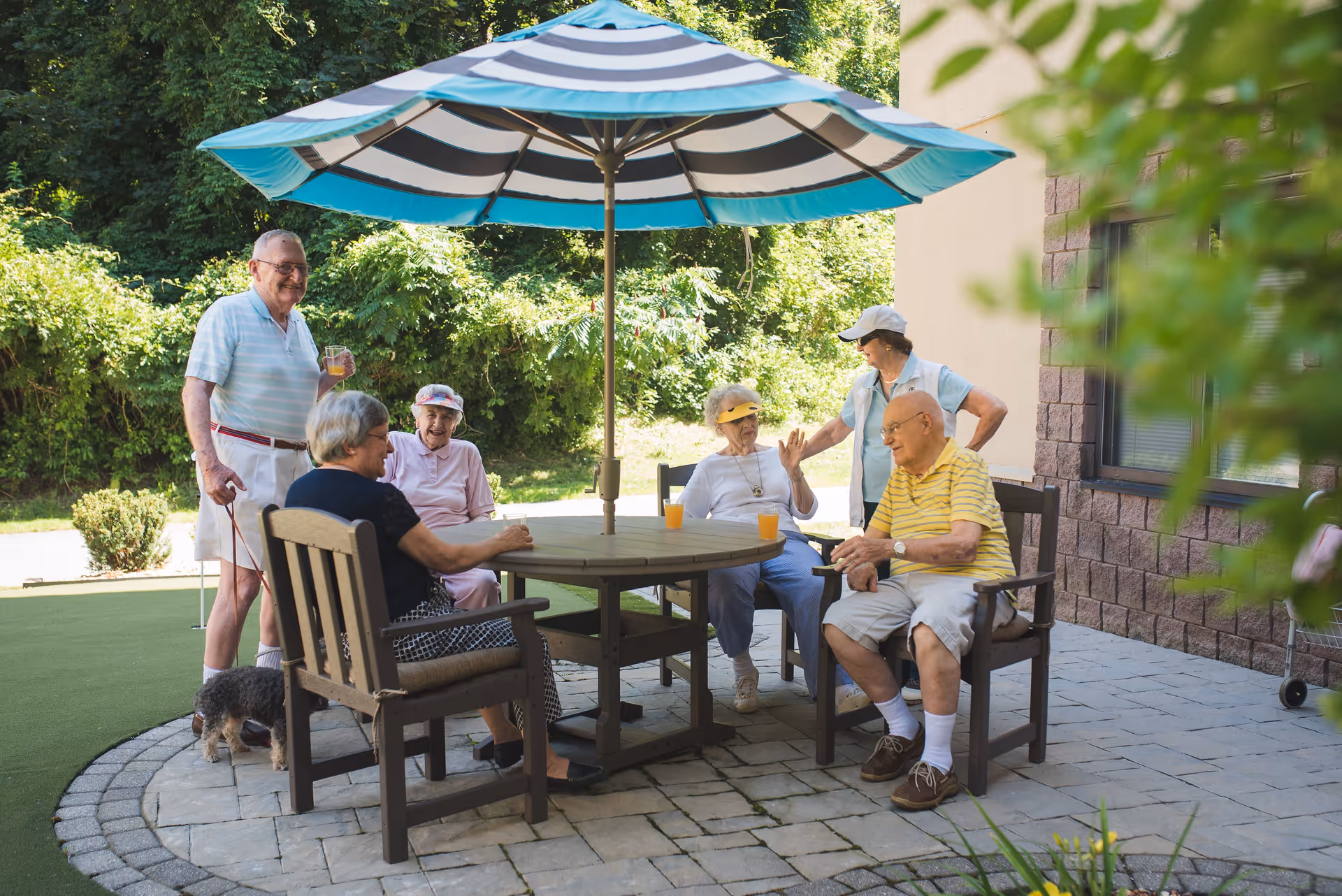 A group of six elderly people sitting and standing around a round outdoor table with a large blue and white striped umbrella. They are enjoying drinks and socializing on a paved patio area next to a building, with greenery and trees in the background. One person is holding a small dog on a leash.