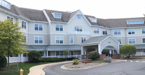 Exterior view of a large, three-story senior living facility building with beige siding and multiple windows. The entrance has a covered driveway with a small landscaped area featuring bushes and flowers. A curved driveway and a fire hydrant are visible in the foreground.
