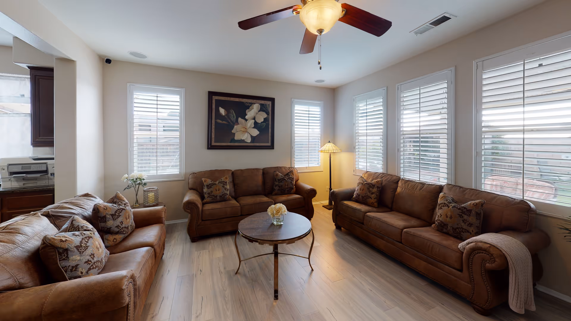A cozy living room with three brown leather sofas arranged around a round wooden coffee table with a small flower vase on it. The room has light-colored wooden flooring, four windows with white blinds, a ceiling fan with a light, a floor lamp in the corner, and a floral painting on the wall.