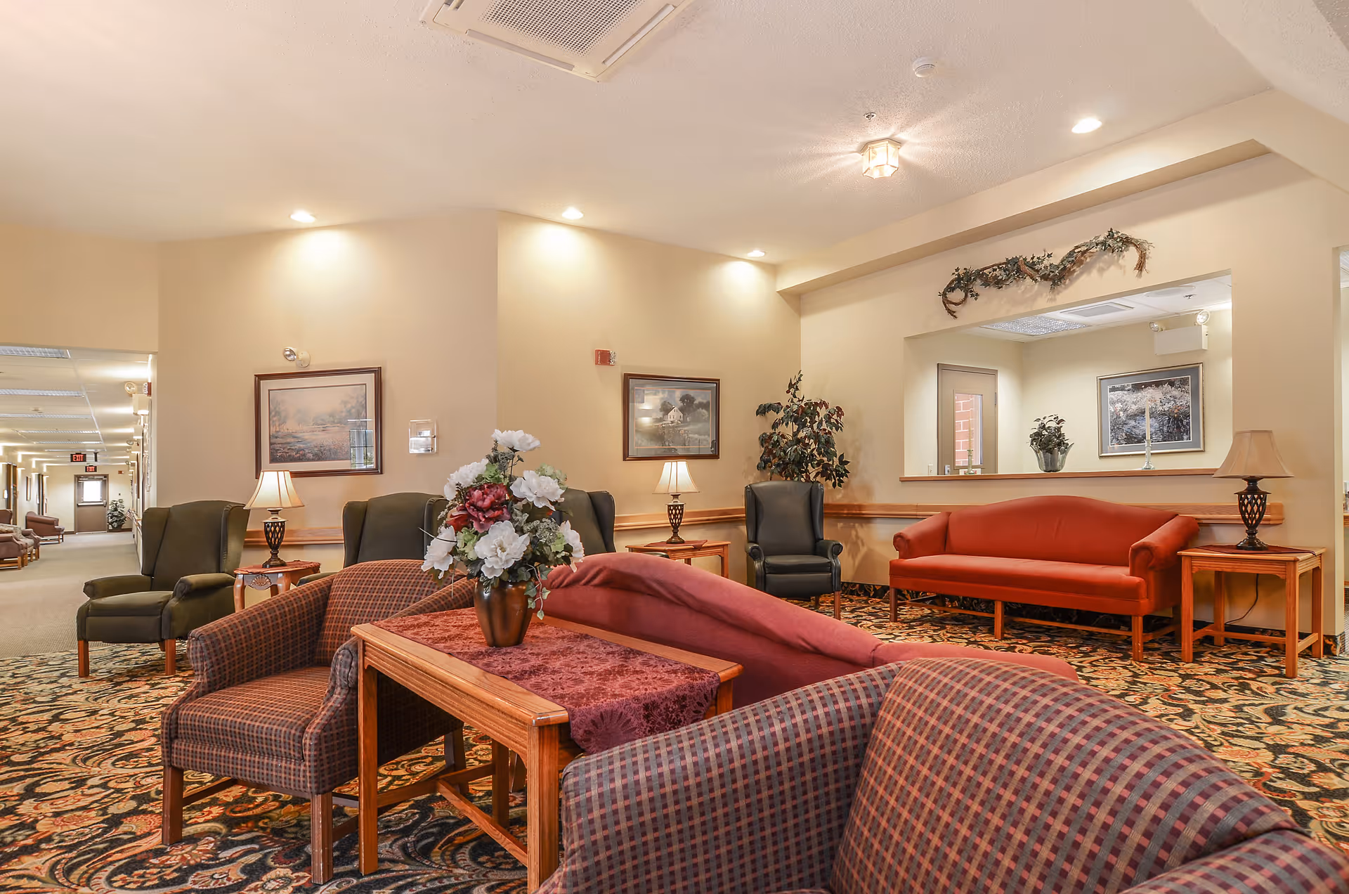 A cozy senior living facility common area with patterned carpet, multiple upholstered chairs and sofas in shades of red and green, wooden side tables with lamps, framed artwork on beige walls, and a floral arrangement on a central wooden table. There is a hallway visible to the left and a reception window on the right wall decorated with greenery.