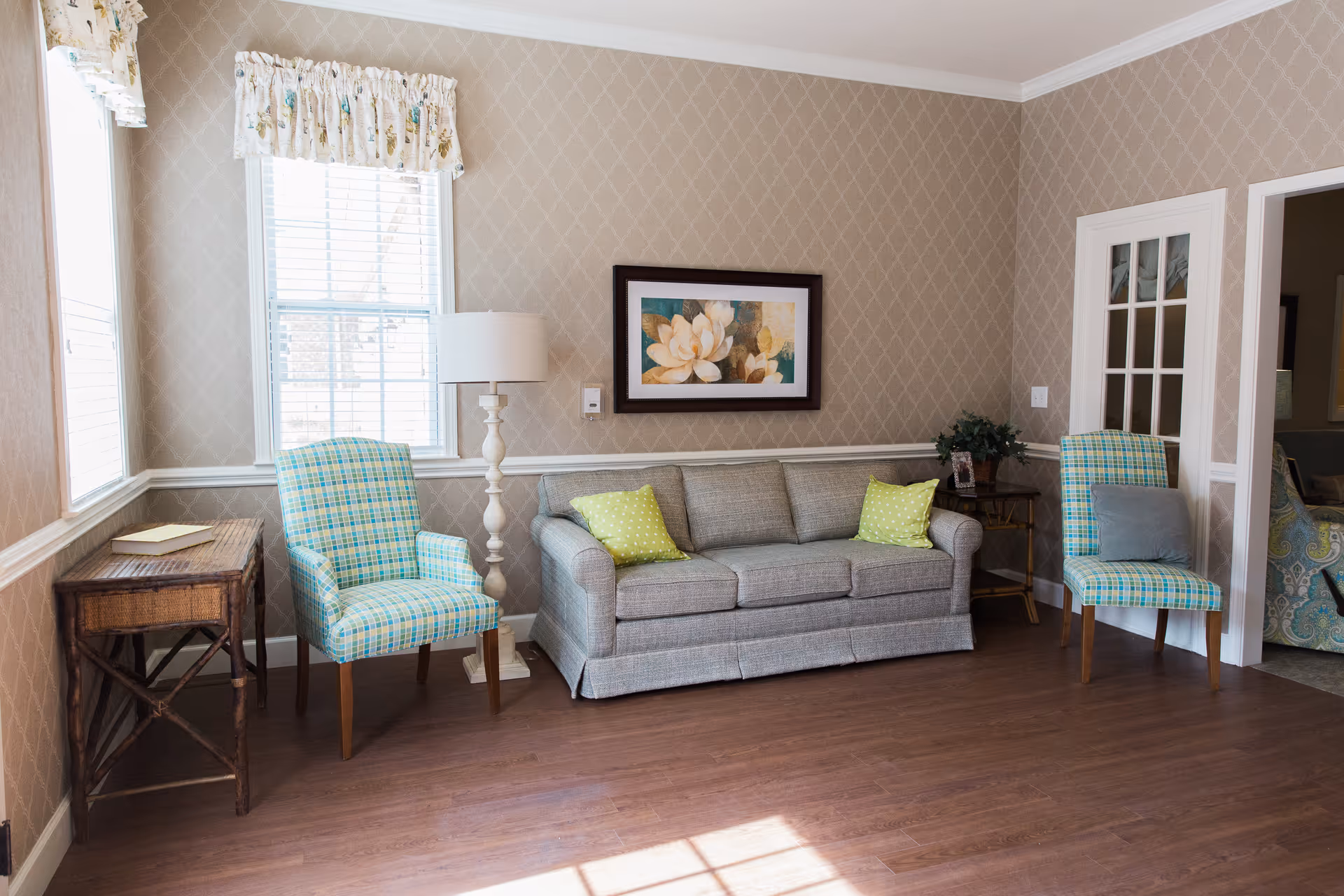 Sunlit living room with a gray sofa, two plaid armchairs, a floor lamp, and framed floral artwork on the wall.