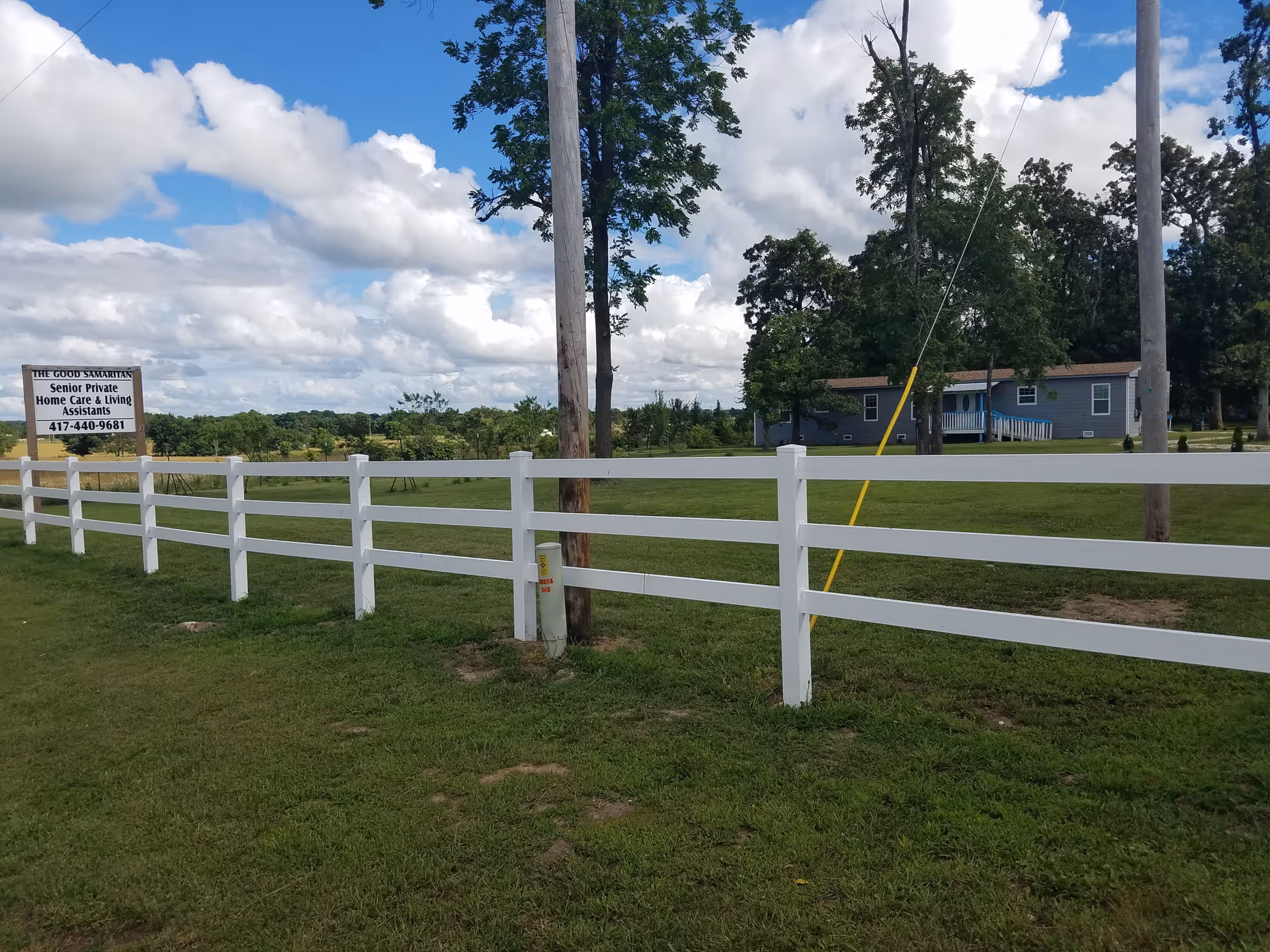 A rural scene showing a white wooden fence running along a grassy area with a sign that reads 'The Good Samaritan Senior Private Home Care & Living Assistants 417-440-9681'. In the background, there is a single-story gray building with a porch, surrounded by trees under a partly cloudy blue sky.