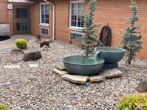 Outdoor courtyard area with a pebble ground cover, two small evergreen trees, a green water fountain with water flowing, a garden hose coiled on the wall, and a brick building with windows and a door in the background.