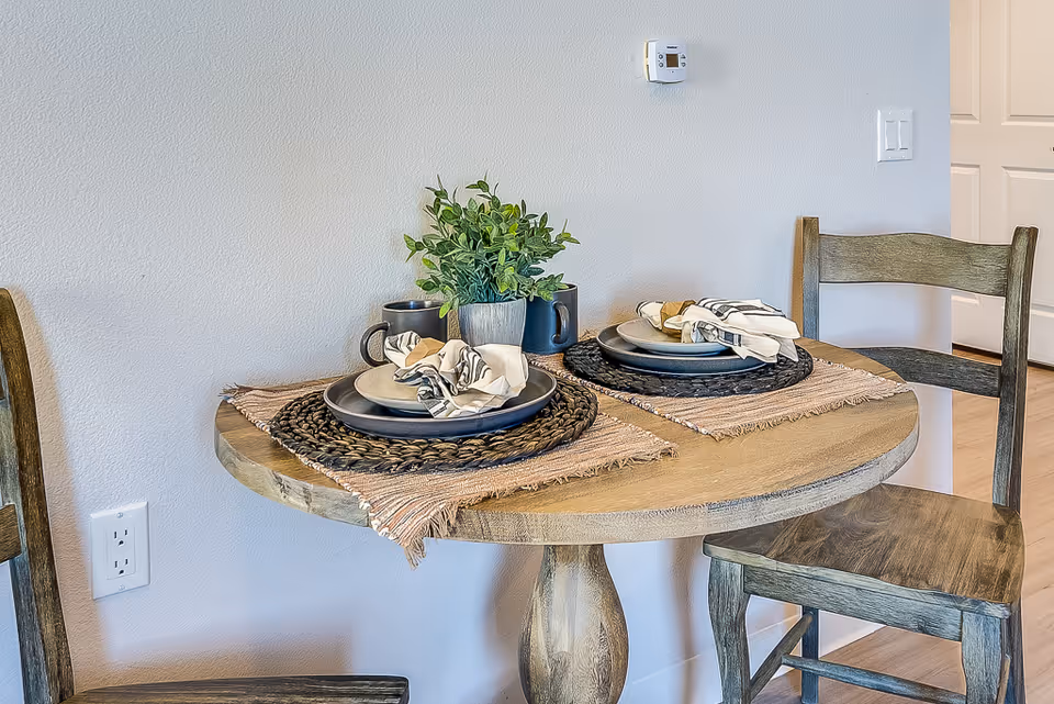 A small round wooden dining table set for two with woven placemats, plates, napkins, mugs, and a small potted plant centerpiece. Two wooden chairs are positioned on either side of the table against a light-colored wall with a thermostat and light switch visible.
