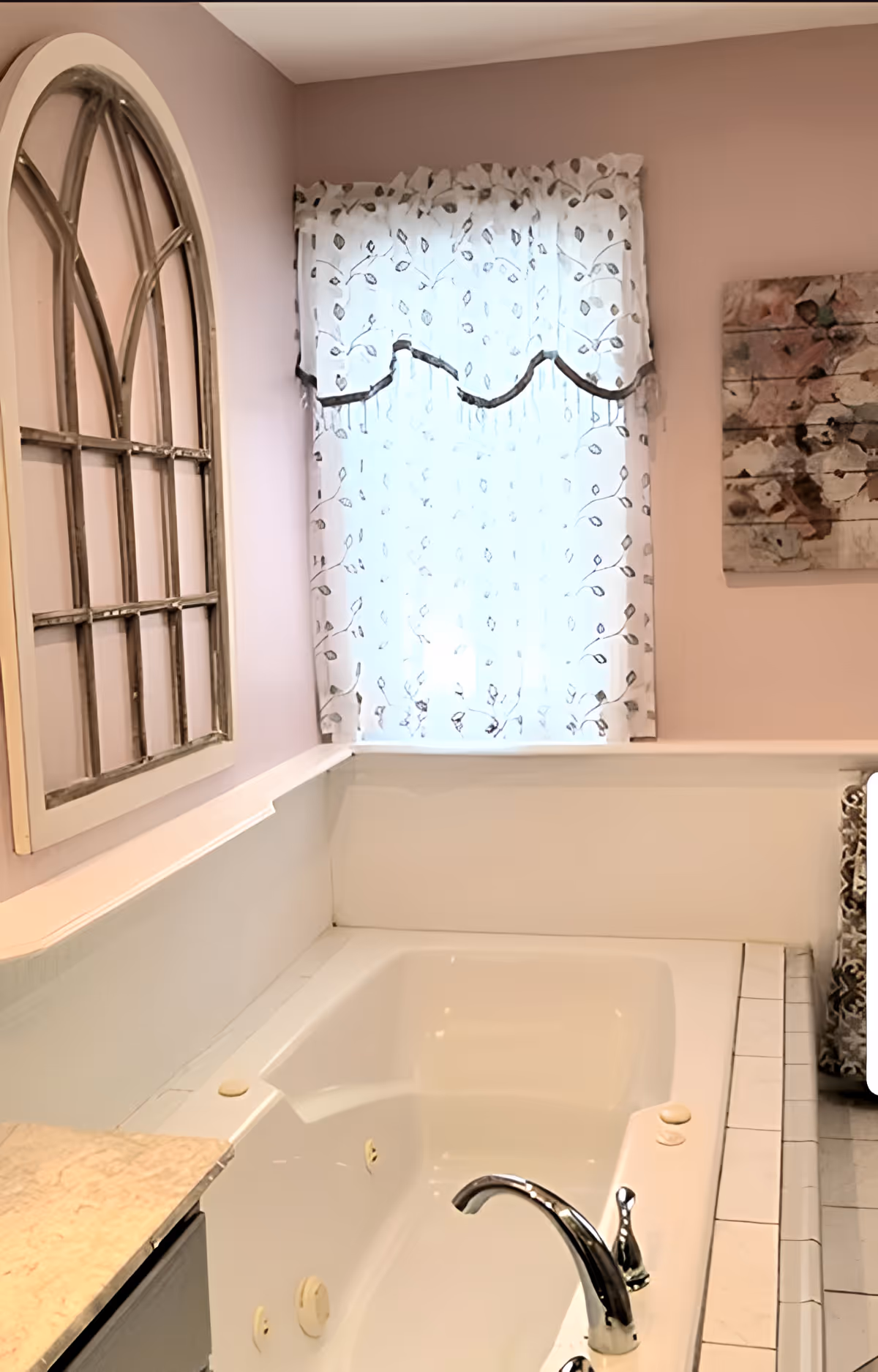 A bathroom featuring a white bathtub with a chrome faucet, a window with sheer white curtains decorated with a leaf pattern, a decorative wall mirror with an arched windowpane design, and a piece of abstract wall art on a light purple wall.