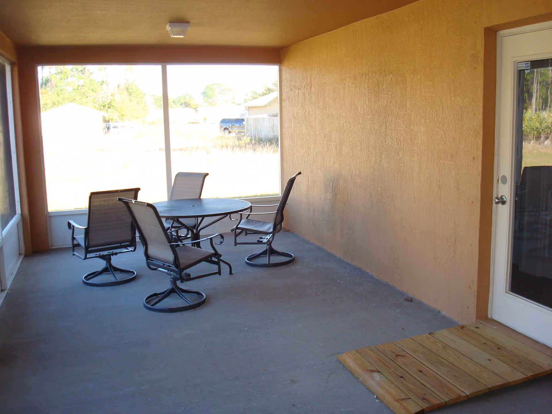A covered outdoor patio area with a round metal table and four swivel chairs with mesh backs and seats. The patio has a concrete floor and is enclosed with large screened windows. There is a wooden ramp leading to a glass door on the right side. Outside, some grass and trees are visible.