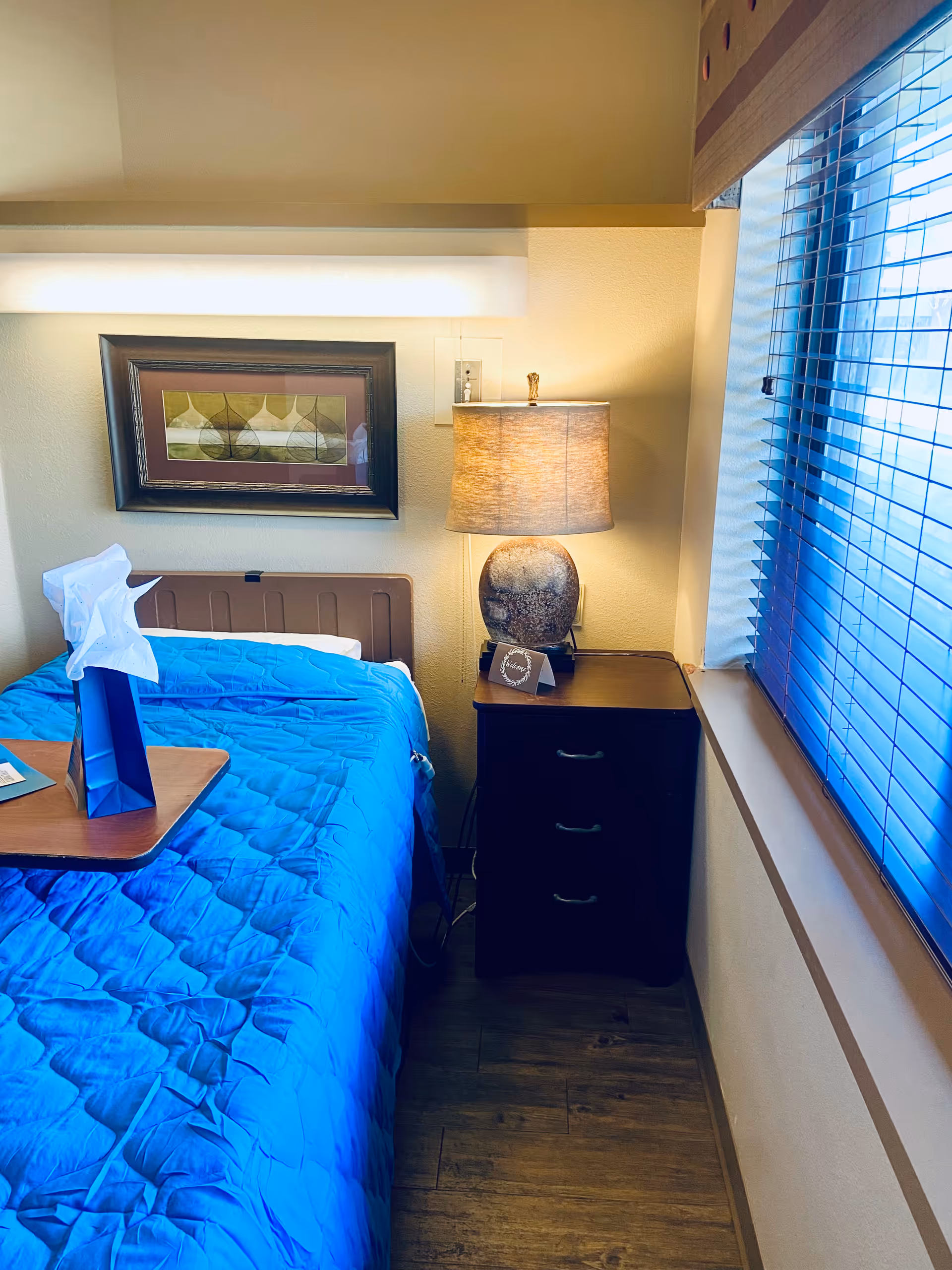 A small bedroom with a single bed covered in a bright blue quilt. Next to the bed is a dark wooden nightstand with three drawers and a lamp on top. Above the bed hangs a framed abstract artwork. A window with blue blinds is on the right side, letting in natural light. A small table with a blue gift bag and tissue paper is positioned over the bed.