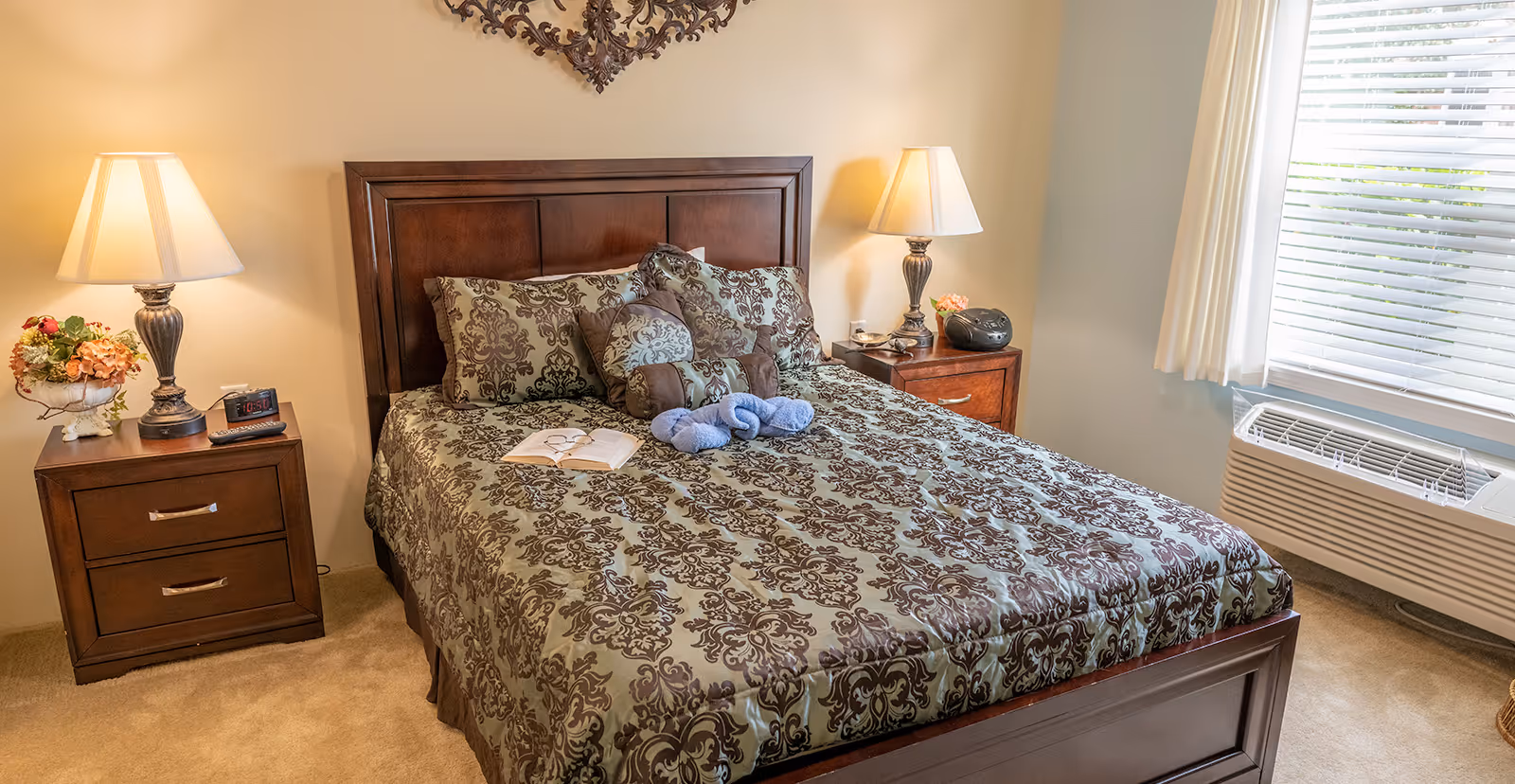 A neatly made bed with a patterned brown and green bedspread and multiple decorative pillows in a bedroom. There are two wooden nightstands on either side of the bed, each with a lamp and decorative items. A window with white blinds and curtains is on the right side, and an air conditioning unit is below the window.