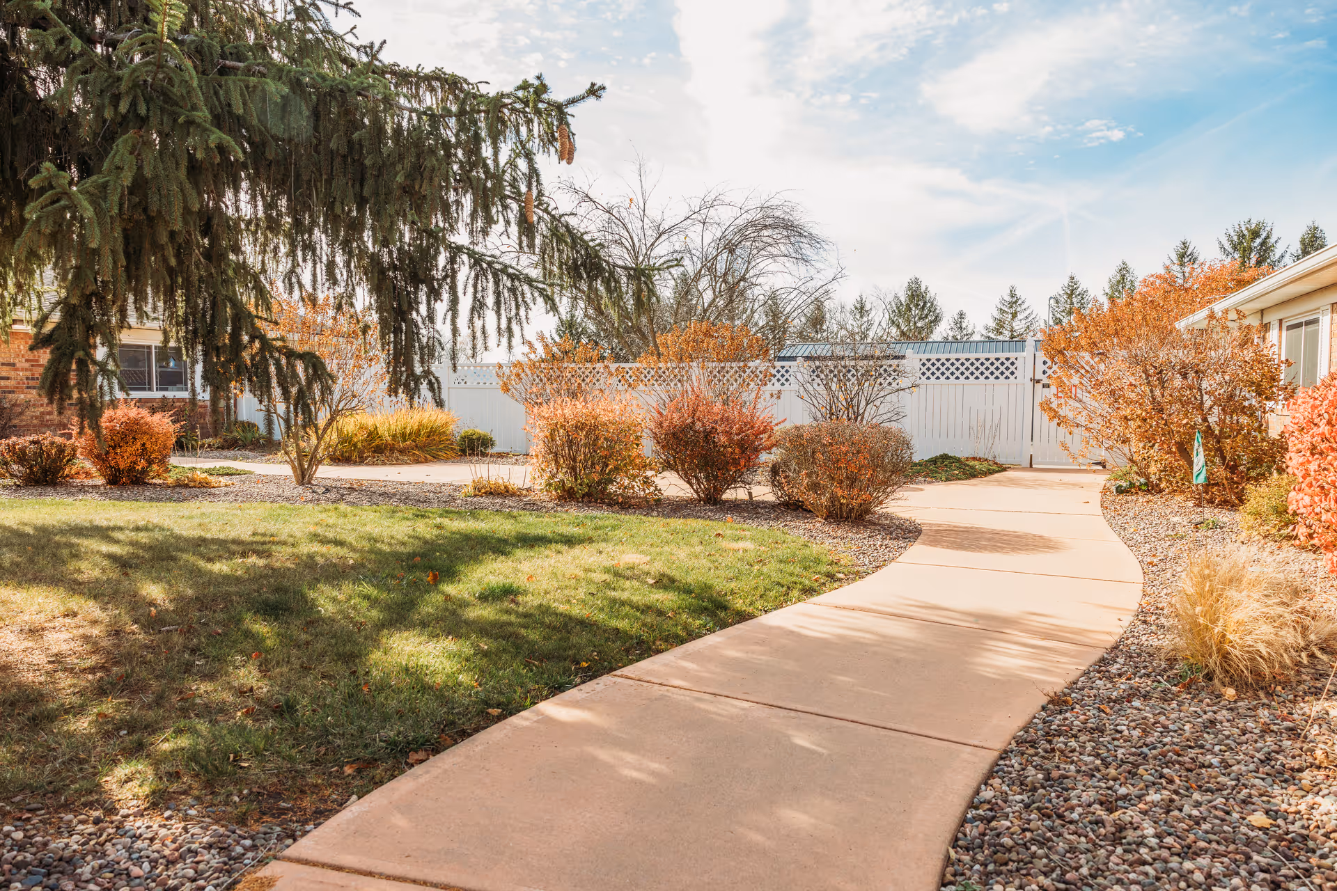 A winding concrete pathway through a landscaped outdoor area with green grass, bushes with autumn-colored leaves, and a large evergreen tree. There is a white fence in the background and parts of a building visible on the left and right sides under a partly cloudy sky.