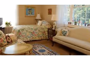 Cozy bedroom with a twin bed in floral bedding, an upholstered sofa, small coffee table and side tables with lamps by a sunlit window.