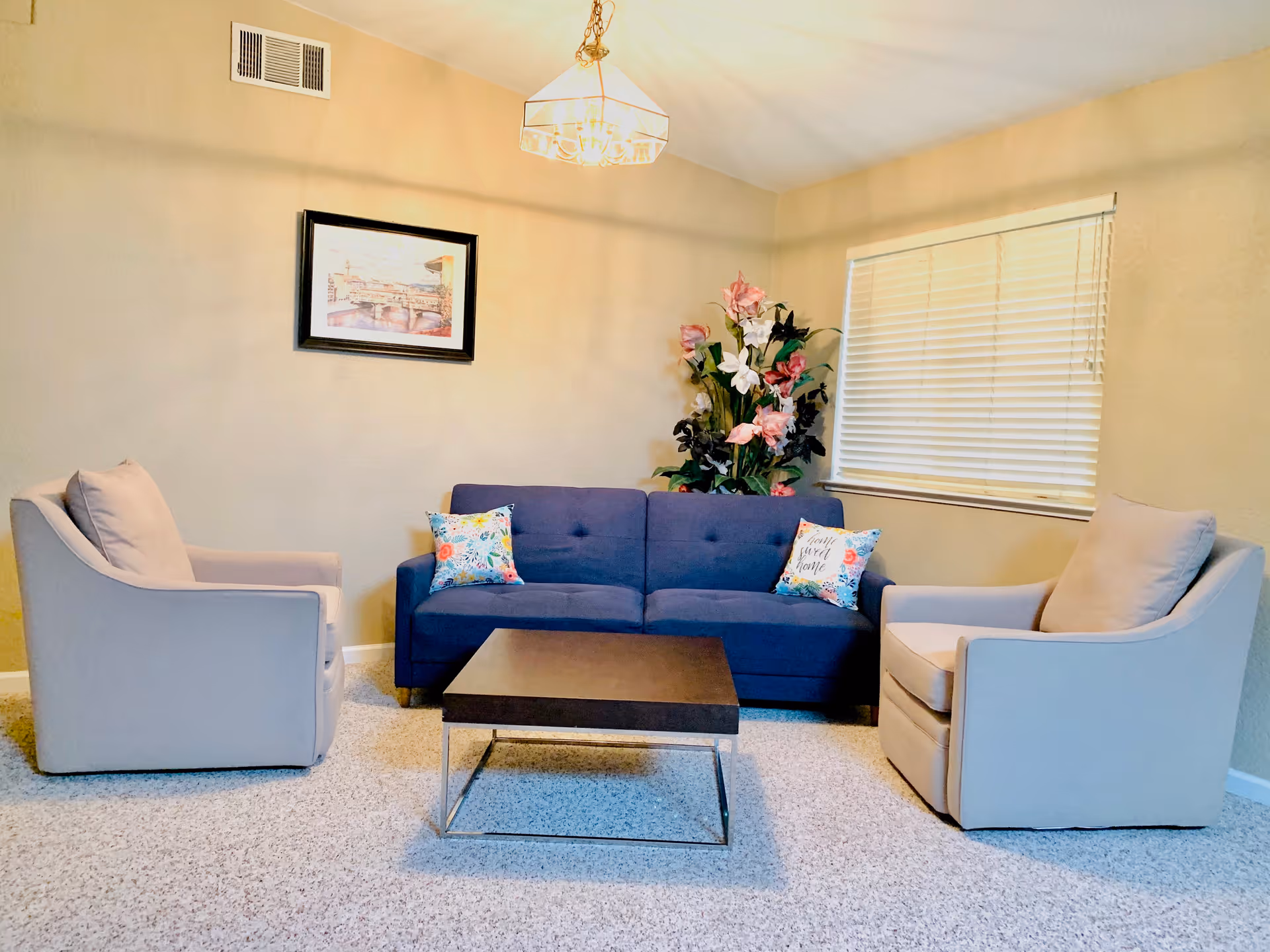 A cozy living room with a blue sofa adorned with two colorful floral pillows, two beige armchairs, a dark wooden coffee table with metal legs, a large floral arrangement in the corner, a framed picture on the wall, and a window with closed blinds.