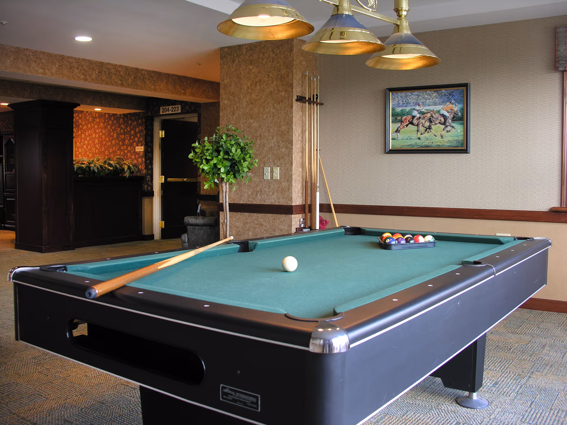 A pool table with a cue stick resting on it and a rack of billiard balls set up on the green felt surface in a room with beige walls, a framed painting of horse riders, a potted plant, and overhead brass light fixtures.