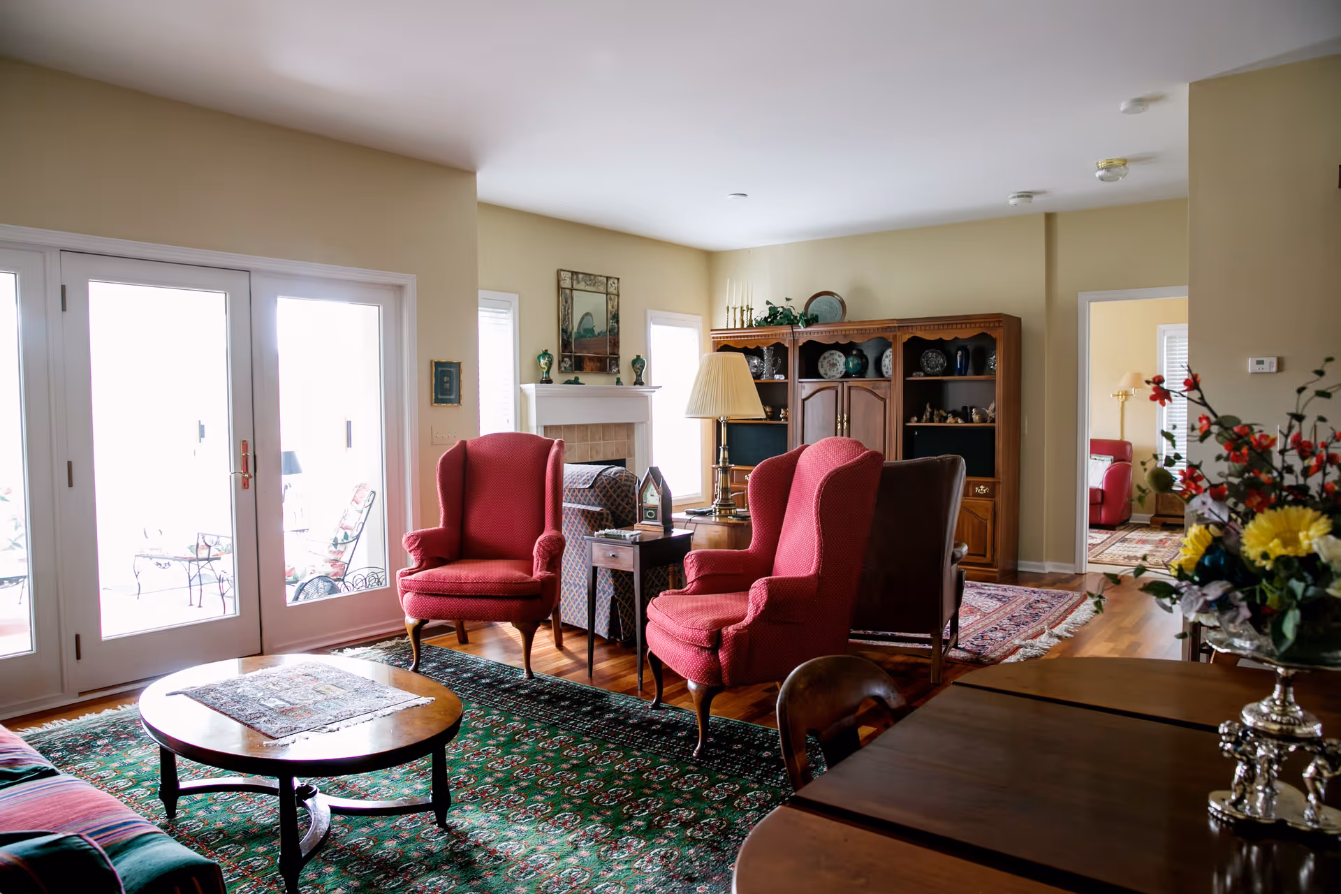 A bright living room with two red wingback chairs facing a round coffee table on a green patterned rug, French doors to a patio, a fireplace, and wooden shelving.