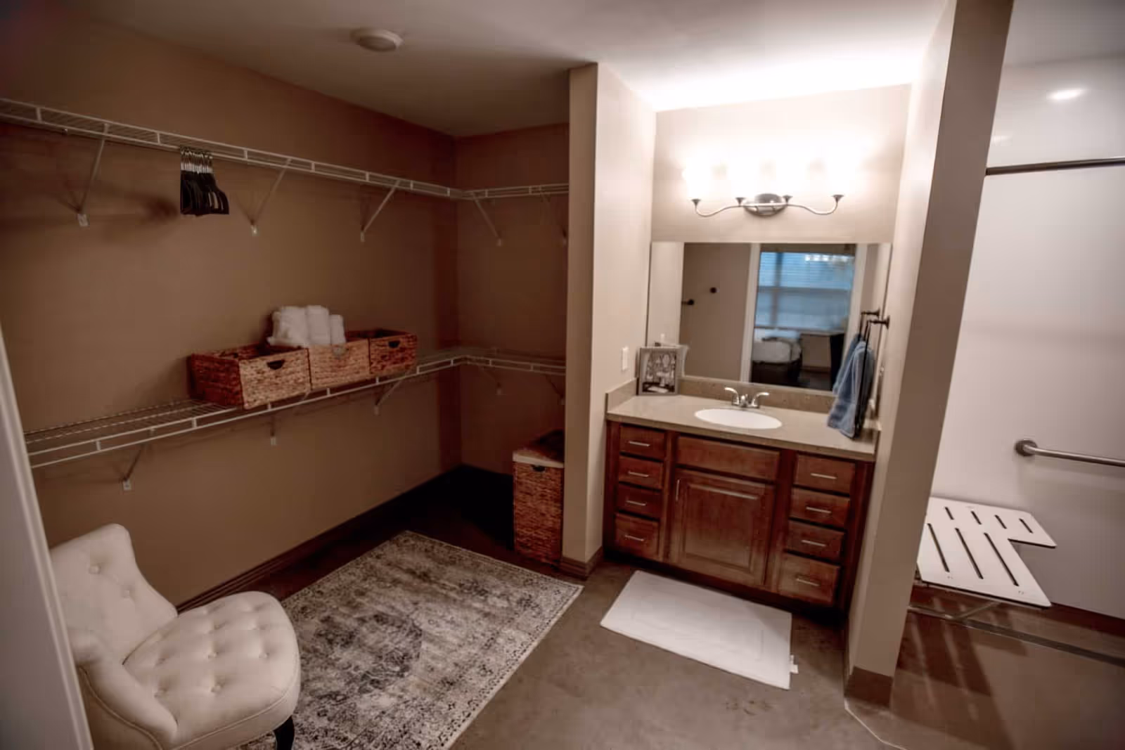 Walk-in bathroom featuring a vanity with mirror and sink, a roll-in shower with bench, and an adjoining open closet with shelving and a tufted chair.
