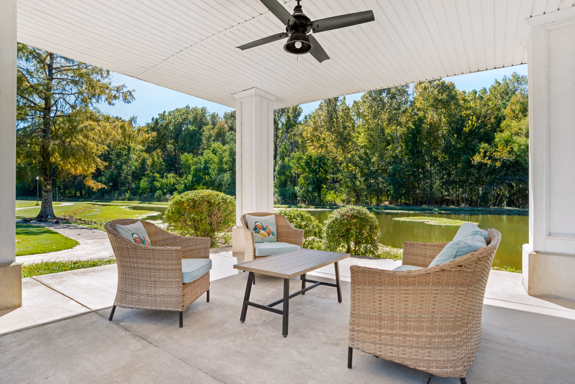 Covered outdoor patio area with four wicker chairs arranged around a rectangular coffee table. The chairs have light blue cushions and decorative pillows. The patio overlooks a pond surrounded by green trees and bushes under a clear blue sky. A ceiling fan is mounted on the white ceiling above the seating area.