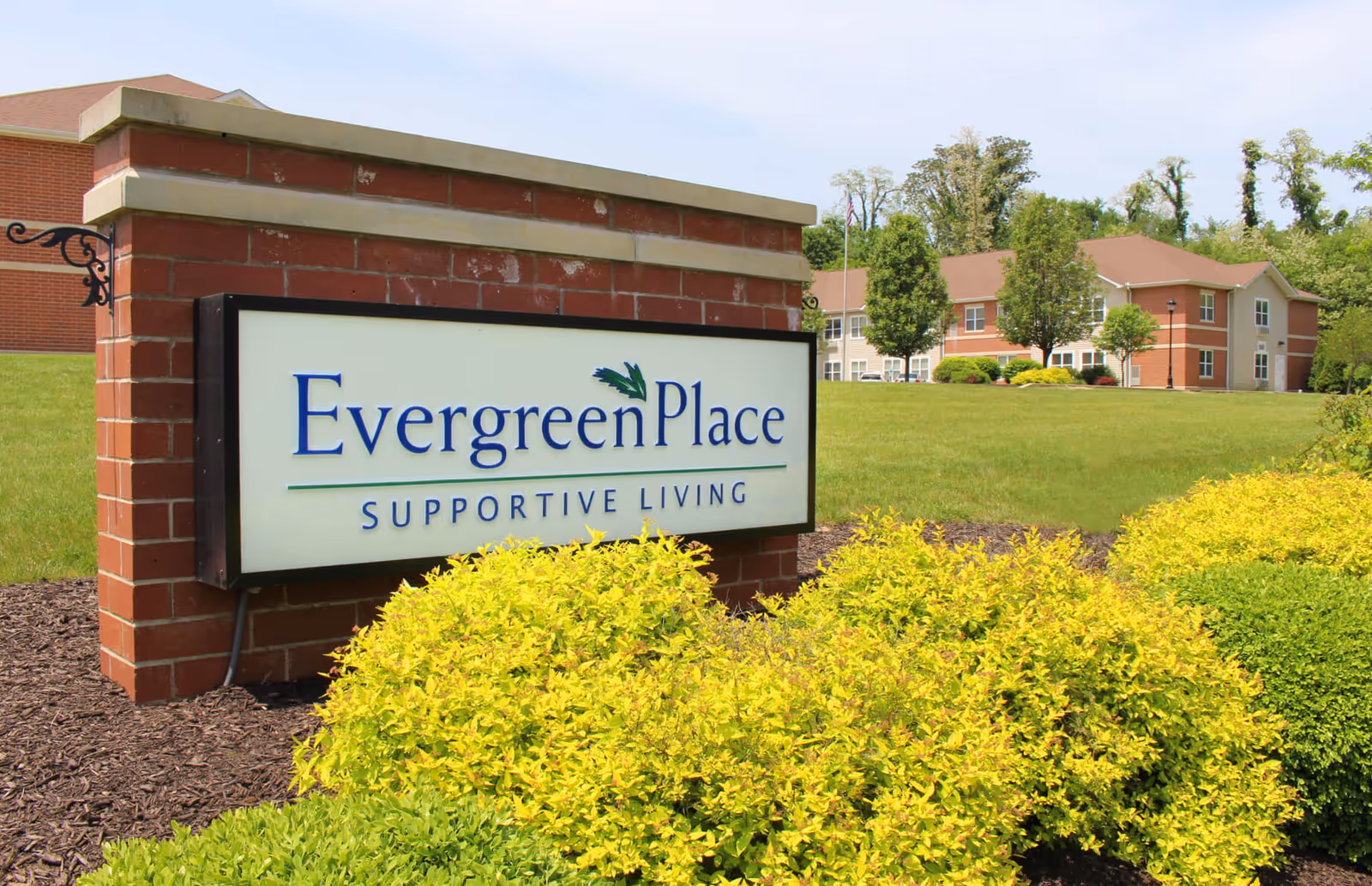 A brick sign for Evergreen Place Supportive Living surrounded by green and yellow bushes with a grassy lawn and residential buildings in the background under a clear sky.