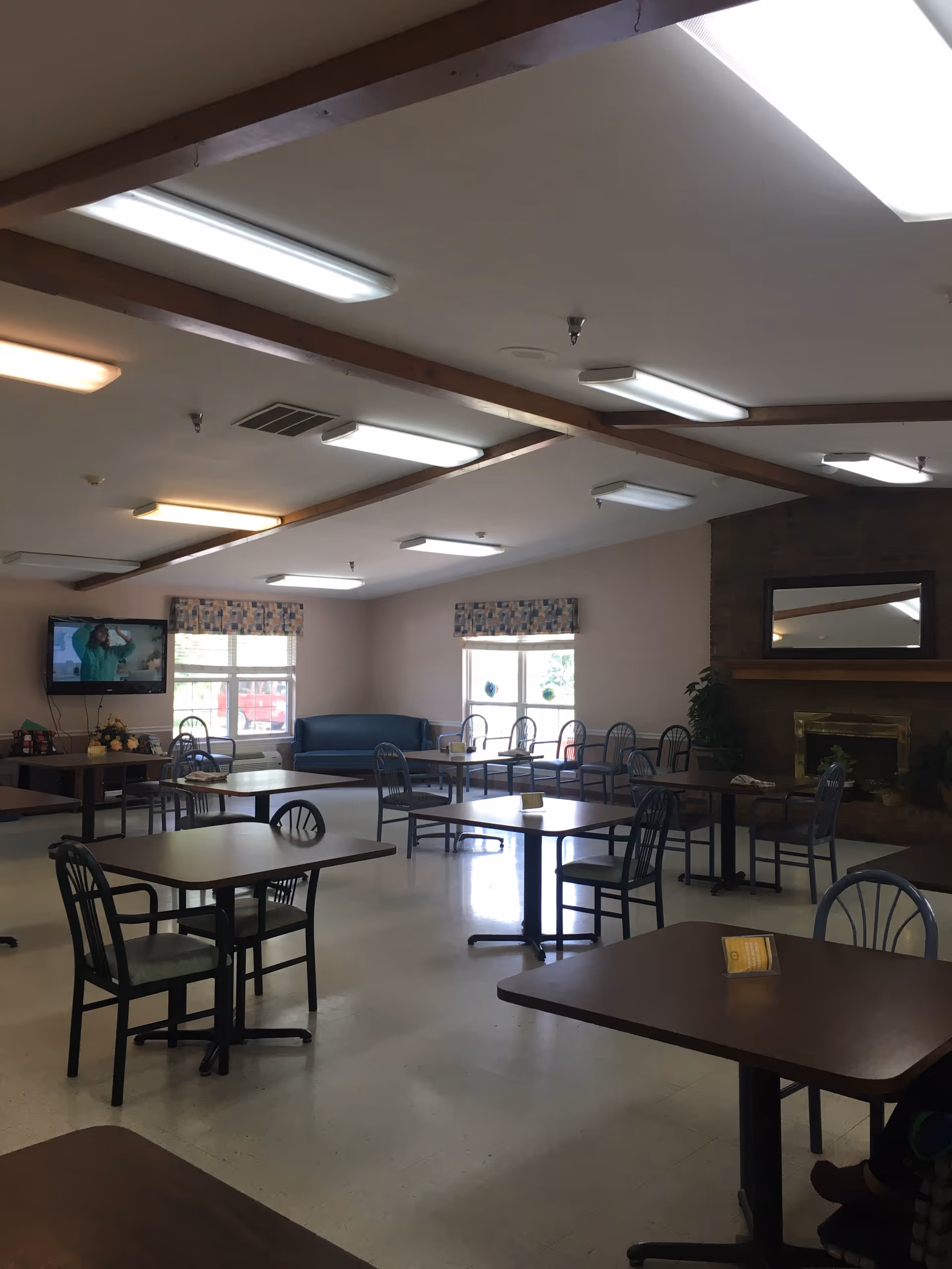 A spacious dining area with multiple tables and chairs arranged neatly. The room has large windows with patterned valances, a wall-mounted television, a blue couch, and a fireplace with a mirror above it. The ceiling has exposed wooden beams and several fluorescent lights.
