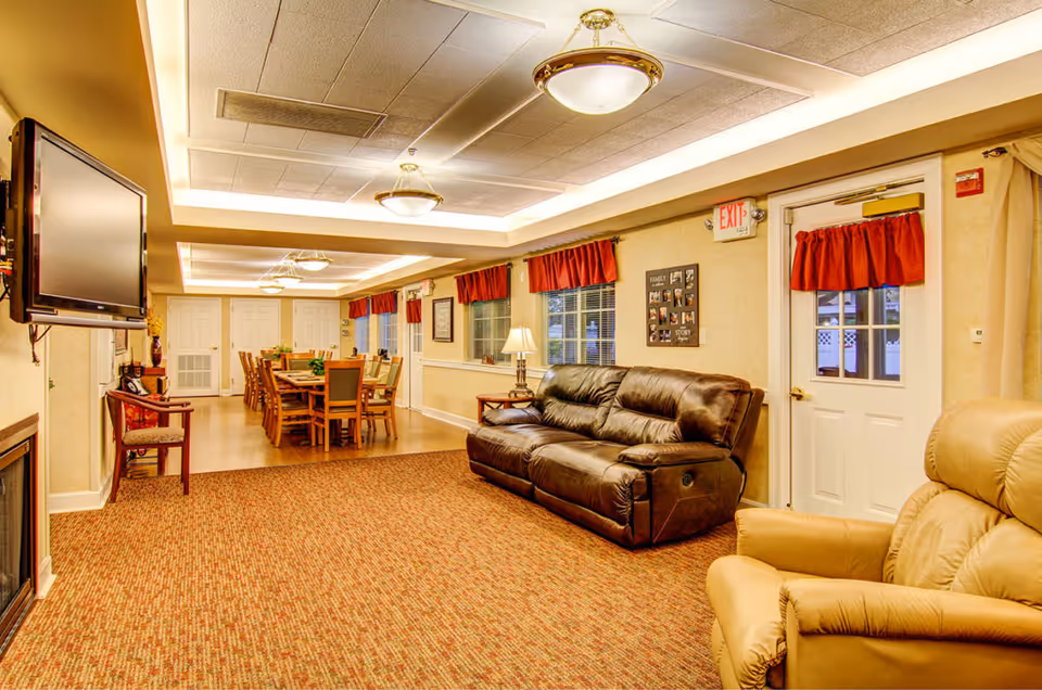 A cozy senior living facility common area featuring a brown leather sofa, a beige recliner chair, a wall-mounted flat-screen TV, and a long wooden dining table with multiple chairs. The room has warm lighting with ceiling fixtures, red curtains on the windows and door, and a carpeted floor.