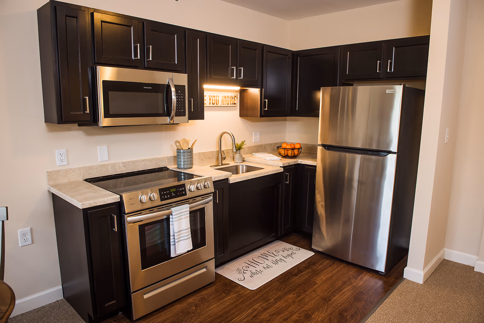A modern kitchen with dark wood cabinets, stainless steel appliances including a microwave, stove, and refrigerator. The countertop is light-colored with a sink and a small plant. There is a decorative sign under the cabinet that says 'YOU MORE' and a floor mat in front of the sink with the phrase 'Home is where our story begins.'