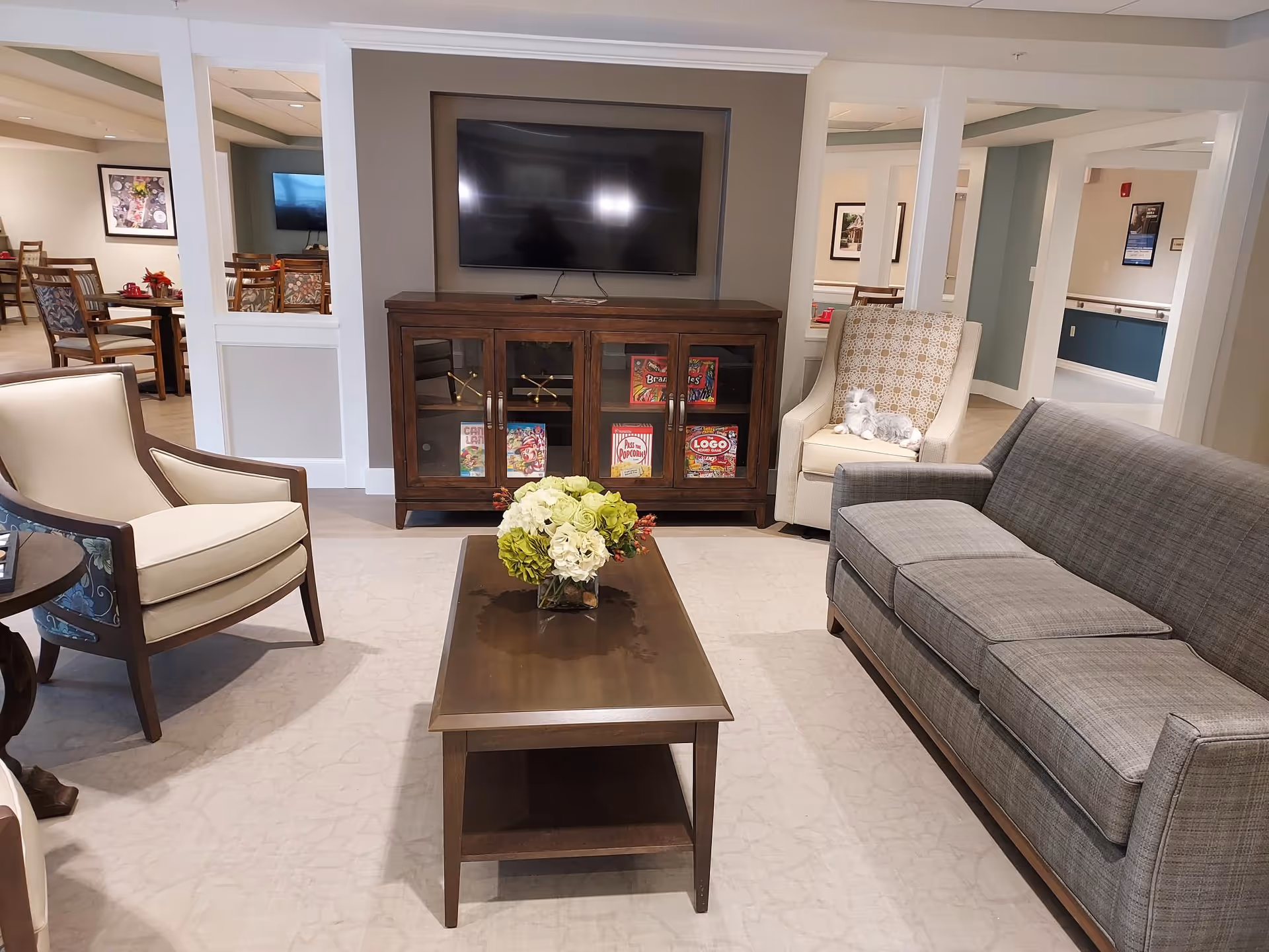 A bright common lounge area with a sofa, armchairs, a coffee table topped with flowers, and a wall-mounted TV over a cabinet.