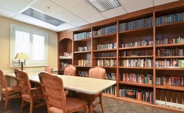 A cozy interior reading room with tall wooden bookshelves along the wall, a rectangular table surrounded by upholstered chairs, and a window with shutters.