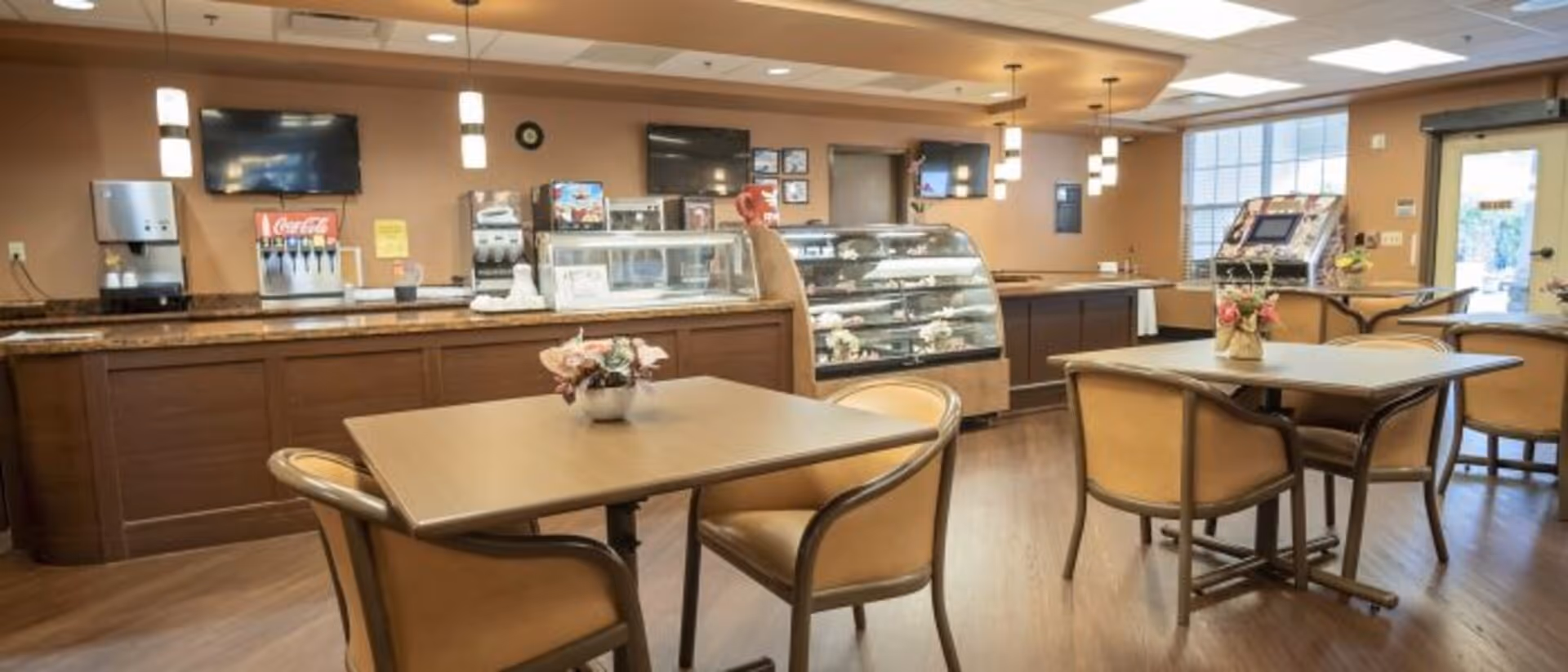 Interior view of a dining area with several tables and chairs. There is a counter along the back wall with a soda fountain, coffee machine, and a glass display case containing food items. The room has warm lighting, beige walls, and large windows allowing natural light to enter.