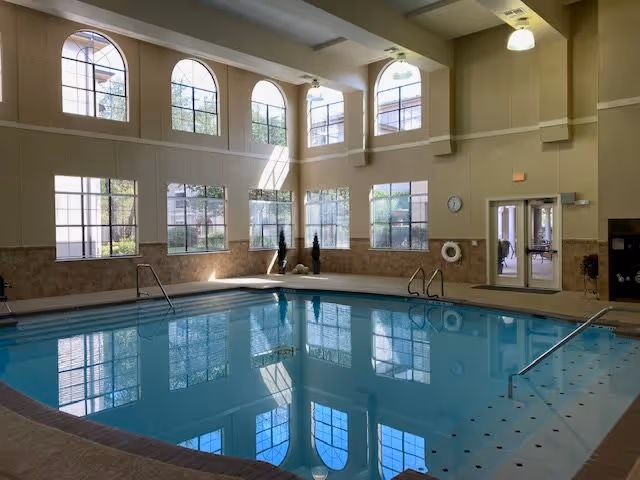 Indoor swimming pool area with large arched windows letting in natural light, beige walls, and tiled pool deck. There are metal handrails for pool entry and exit, a clock on the wall, and a life preserver hanging nearby.