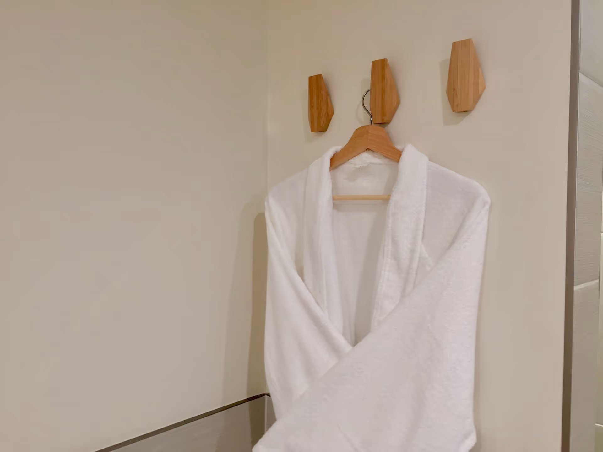 A white bathrobe hanging on a wooden hanger attached to a wooden wall hook on a beige wall in a bathroom.