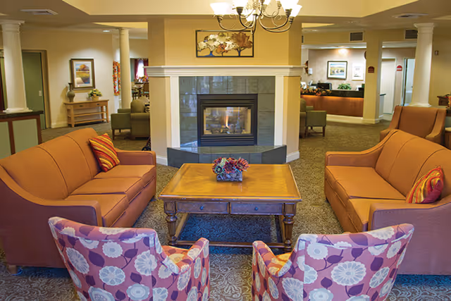 A cozy living room area with two orange sofas facing each other, two floral patterned armchairs, a wooden coffee table with a floral centerpiece, and a fireplace in the background. The room has warm lighting and a carpeted floor, with a chandelier hanging from the ceiling.