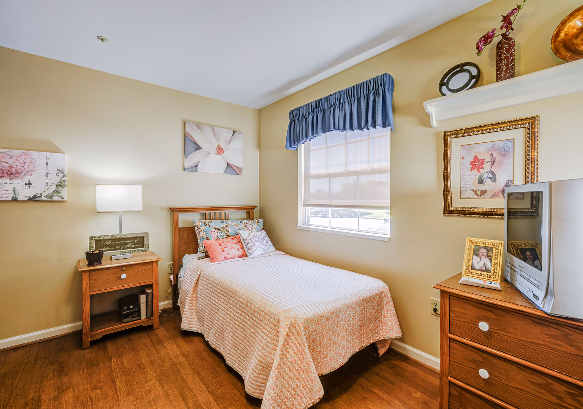 A cozy bedroom with a single bed covered in a light pink quilt and multiple decorative pillows. There is a wooden nightstand with a lamp and a small plant next to the bed. A window with a blue valance and a white shade lets in natural light. On the opposite side, there is a wooden dresser with a small TV and framed photos. The walls are decorated with floral artwork and a shelf holding decorative items.