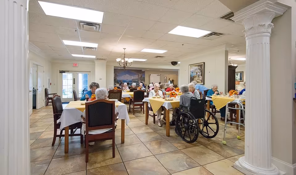 A dining room in a senior living facility with elderly residents seated at tables covered with white and yellow tablecloths. Some residents are using wheelchairs and walkers. The room has tiled floors, white walls, decorative columns, and ceiling lights. Paintings and a chandelier are visible in the background.
