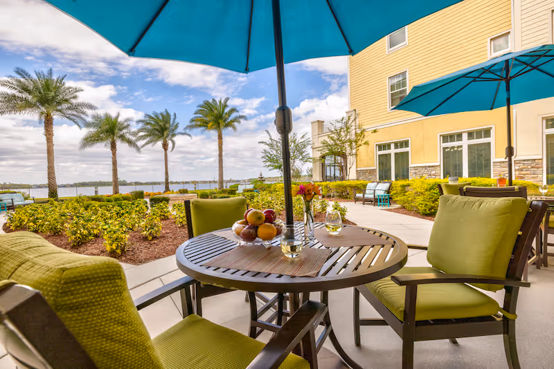 Sunny outdoor patio at Sonata Windermere with a round table, green-cushioned chairs and blue umbrellas overlooking palm trees and a waterfront.