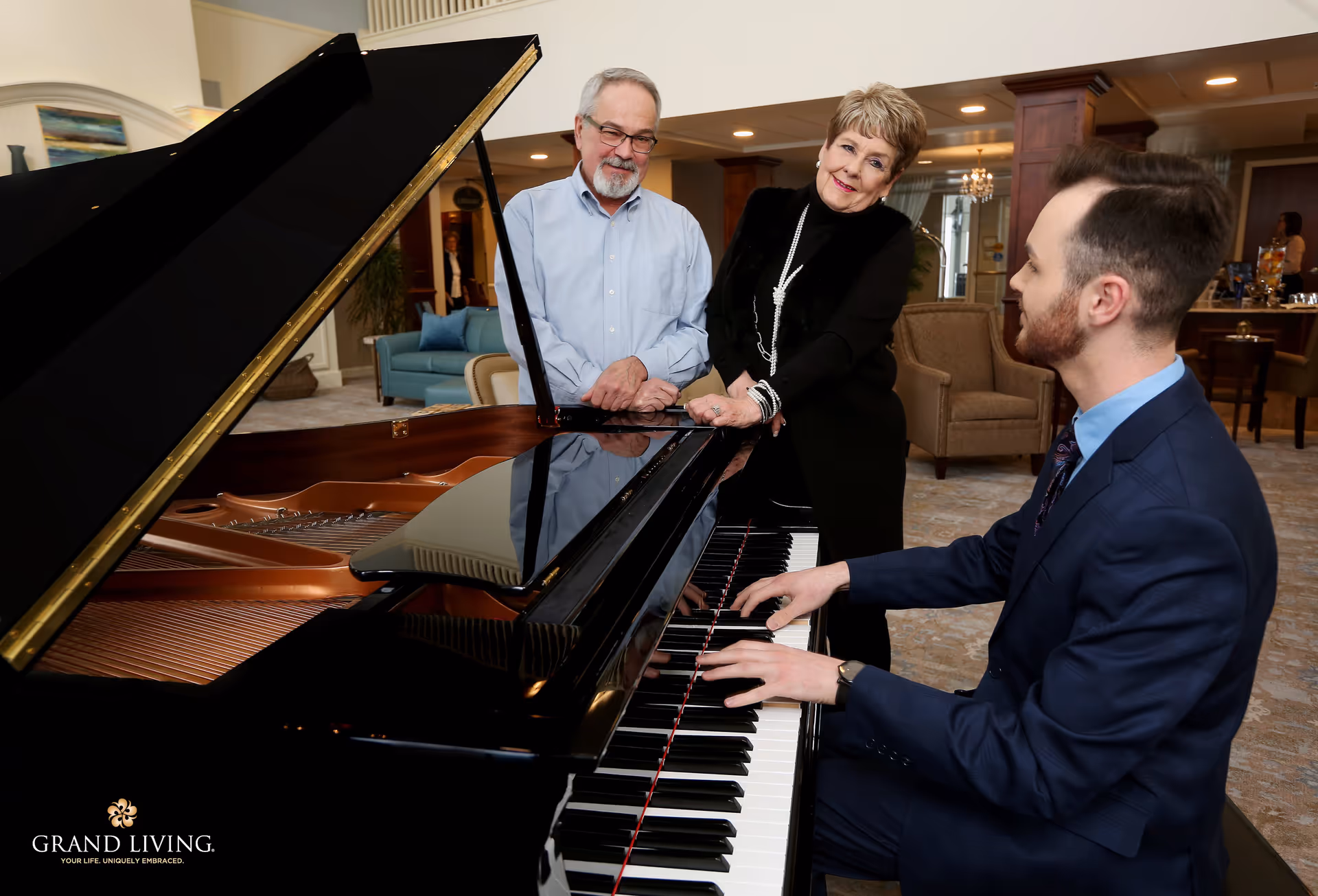 A young man plays a grand piano as an older couple listens in a furnished common room.