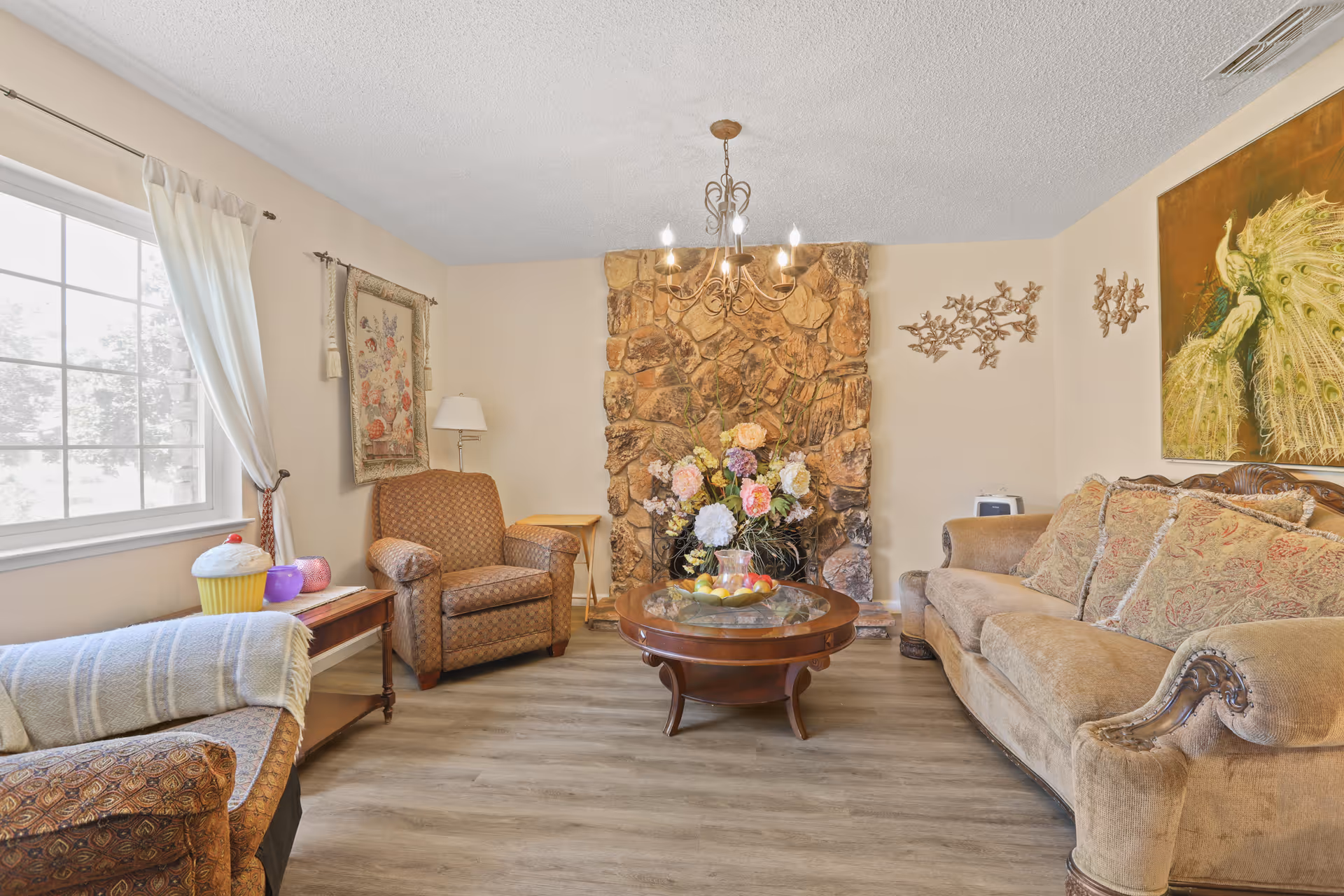 Cozy living room with a stone fireplace, chandelier, sofa and armchairs around a round coffee table topped with a floral arrangement.