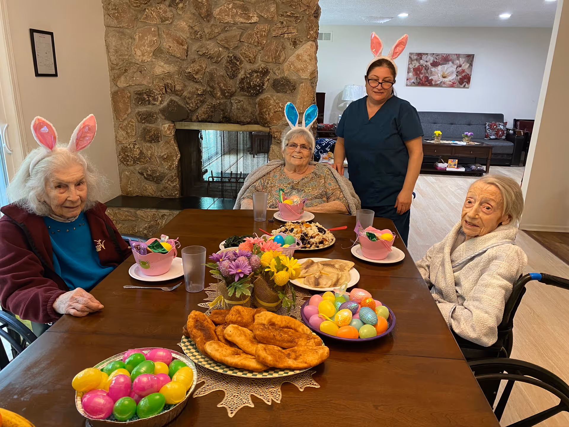 Three elderly women wearing bunny ears sitting around a wooden table with Easter-themed decorations and food, including colorful plastic eggs and pastries, with a caregiver standing behind them in a cozy living room with a stone fireplace.