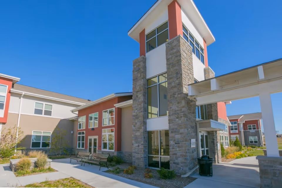 Exterior view of The Healthcare Resort Of Topeka building with stone pillars, large windows, and a covered entrance. There are benches and landscaped plants along the walkway under a clear blue sky.