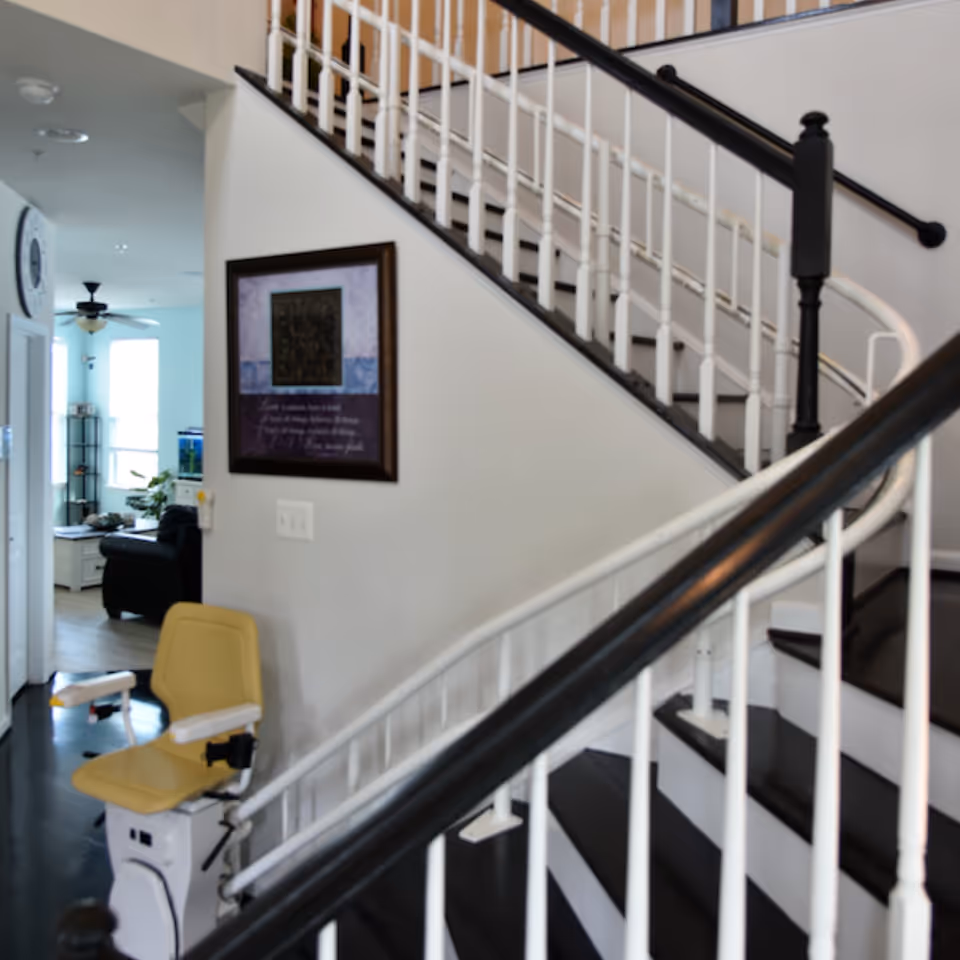 Interior view of a staircase with a yellow stairlift and black-and-white handrails, a framed picture on the wall, and a living area visible in the background.