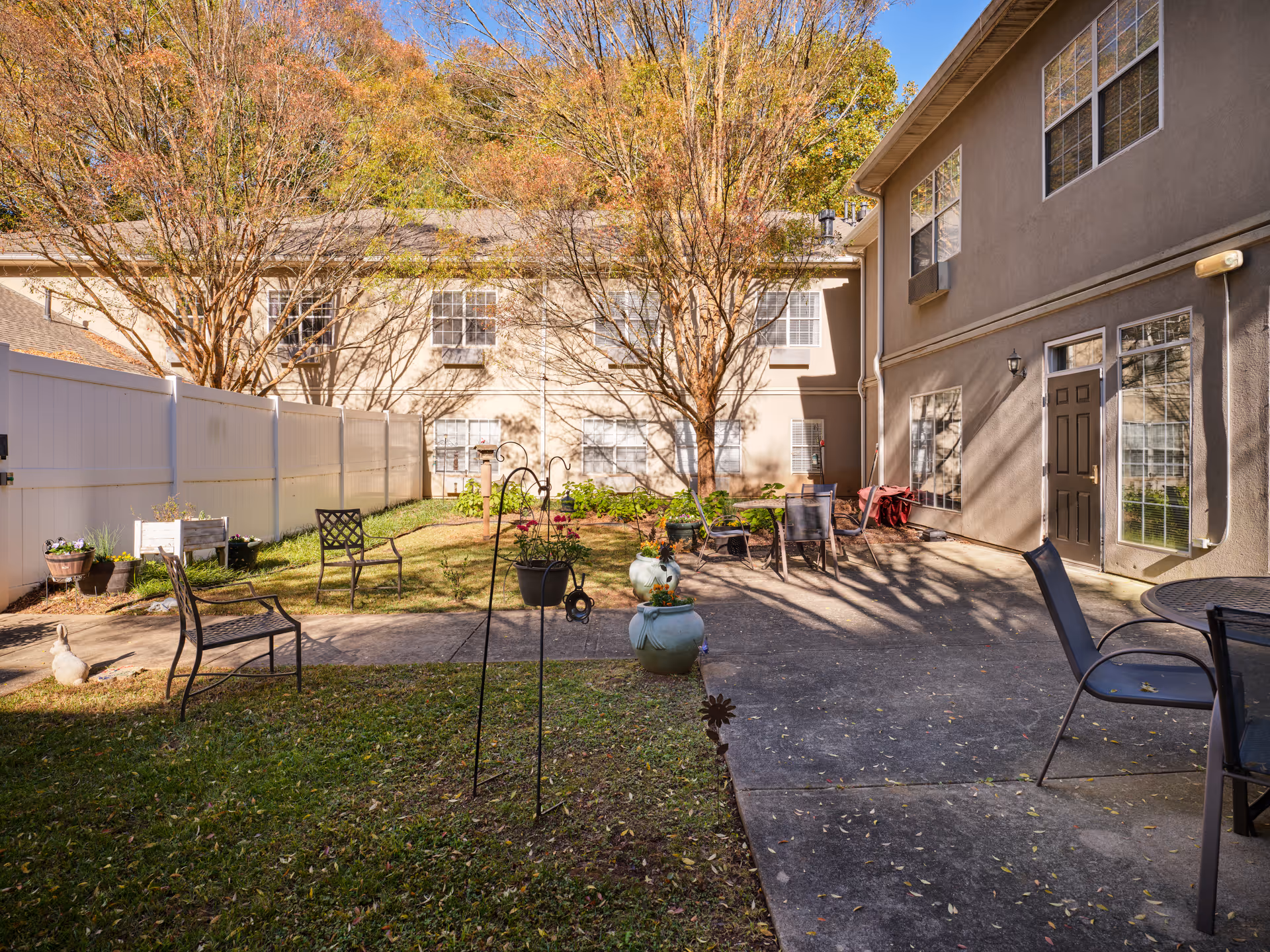 Outdoor courtyard area at Greenwood Place Assisted Living and Memory Care featuring a paved patio with metal chairs and tables, surrounded by grass, potted plants, and leafless trees. The courtyard is enclosed by a white fence and adjacent to a beige two-story building with multiple windows and doors.