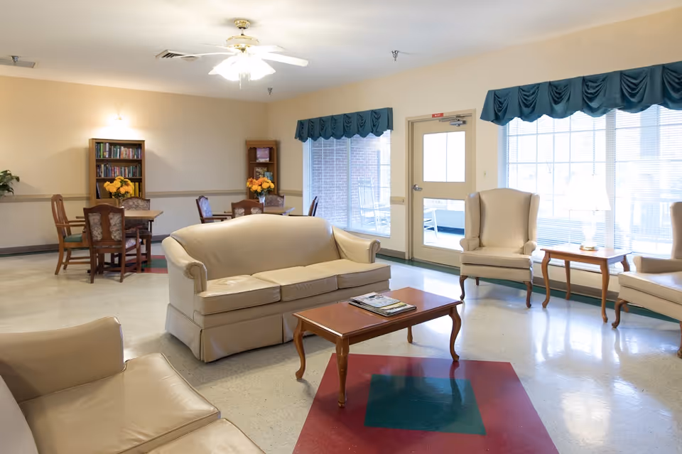 Bright senior living lounge with beige sofas, chairs, tables, bookshelves, and large windows.