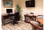 Small interior common room with a desk and computer, wall-mounted TV, table and chairs, potted plant, and framed picture.