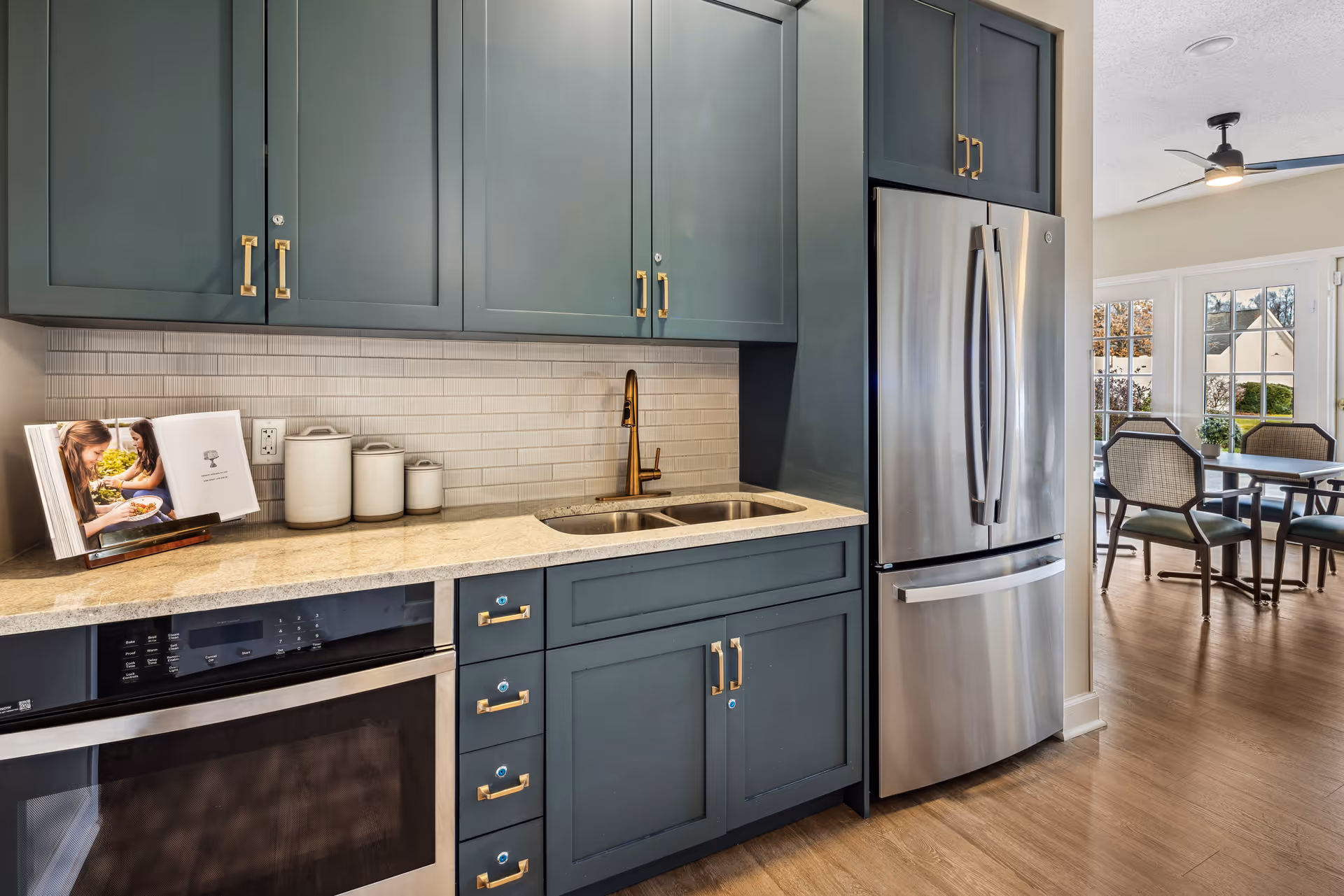 Modern kitchen area with blue-gray cabinets, a stainless steel refrigerator, a built-in oven, a double sink with a bronze faucet, and a countertop with three white canisters and an open cookbook. In the background, there is a dining area with a table, chairs, large windows, and a ceiling fan.