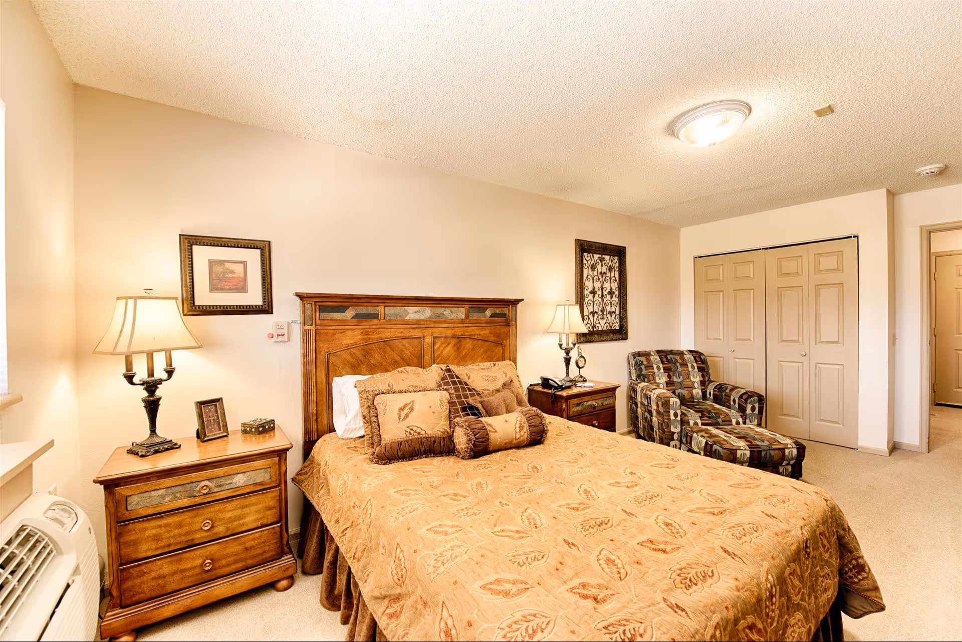 A warmly lit bedroom featuring a wooden bed with a patterned beige and brown bedspread and multiple decorative pillows. On either side of the bed are wooden nightstands with lamps, a framed picture on the left wall, and a decorative wall hanging on the right. There is a patterned armchair with an ottoman near a set of closed double doors, and a beige carpet covers the floor.