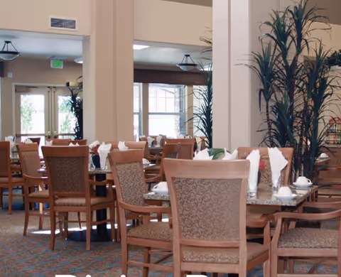 A dining room with multiple tables and chairs arranged neatly. Each table is set with white napkins, cups, and plates. There are large windows and glass doors in the background letting in natural light, and tall green plants are placed near the walls.