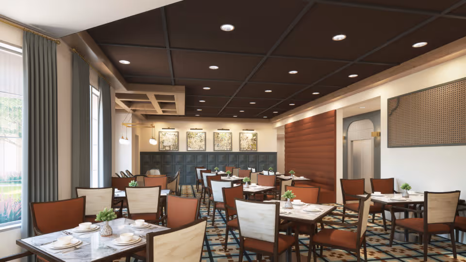 A spacious dining room with multiple tables set with white plates, cups, and small green plants as centerpieces. The room features large windows with gray curtains, a patterned floor, and a ceiling with recessed lighting. The walls are decorated with framed artwork and a textured panel.