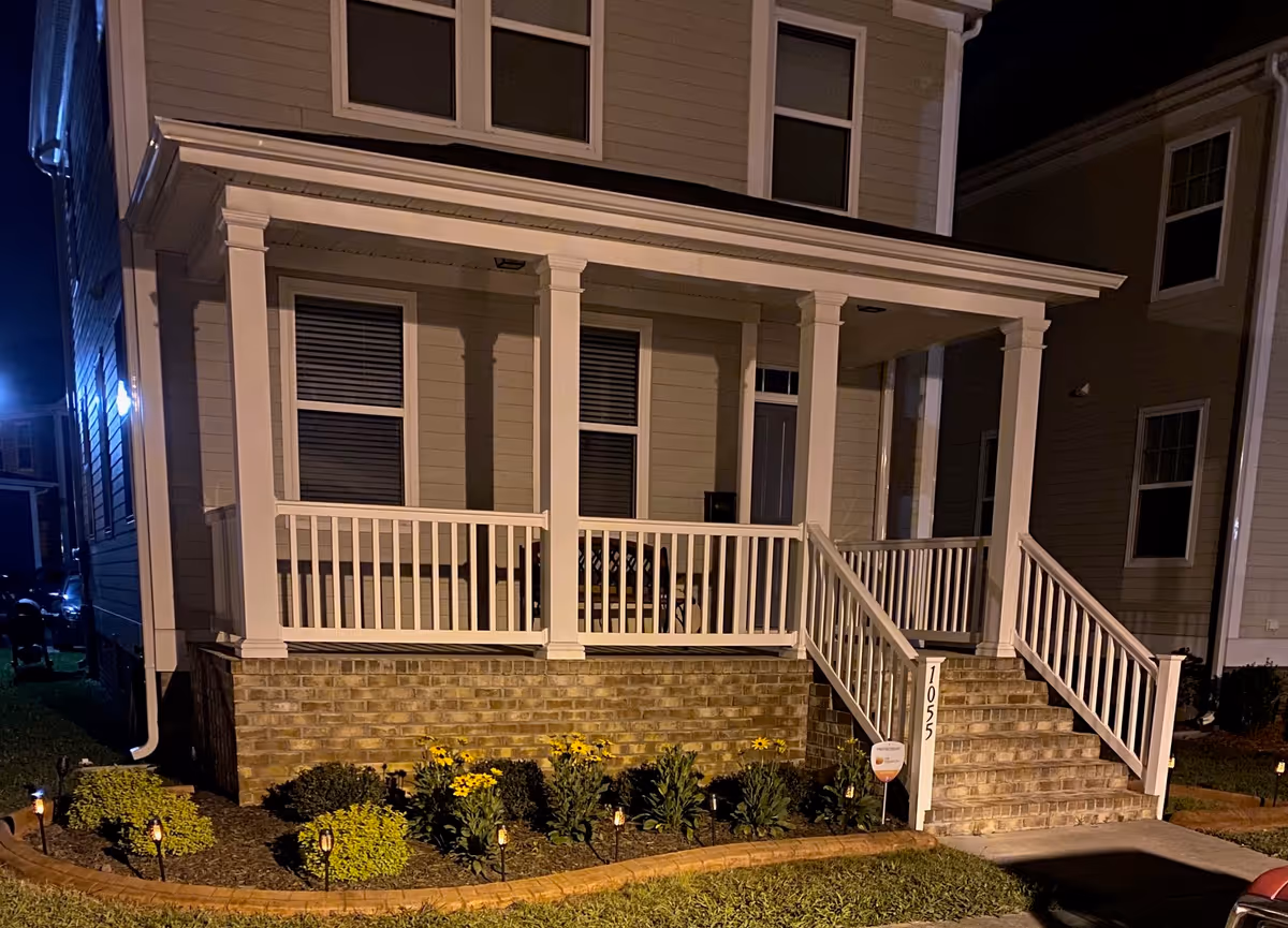 A nighttime view of the front porch of a house with white railings and columns, brick steps, and a small garden with yellow flowers and bushes along the front. The house number 1052 is visible on a post beside the steps.