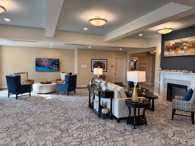 A spacious and well-lit living room area in a senior living facility with patterned carpet flooring, multiple seating arrangements including armchairs and sofas, a round ottoman, a fireplace with a decorative mantle, and framed artwork on the walls. The room has ceiling lights and table lamps providing warm lighting.