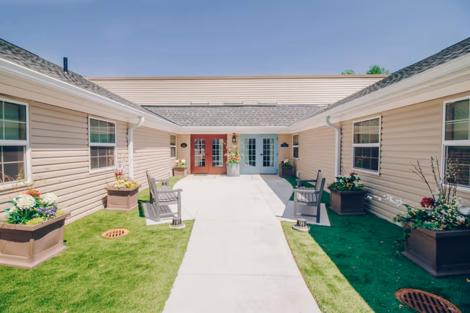 Outdoor courtyard area of a senior living facility with beige siding buildings on both sides, green grass, flower planters, benches, and two sets of double doors, one red and one light blue, under a clear blue sky.