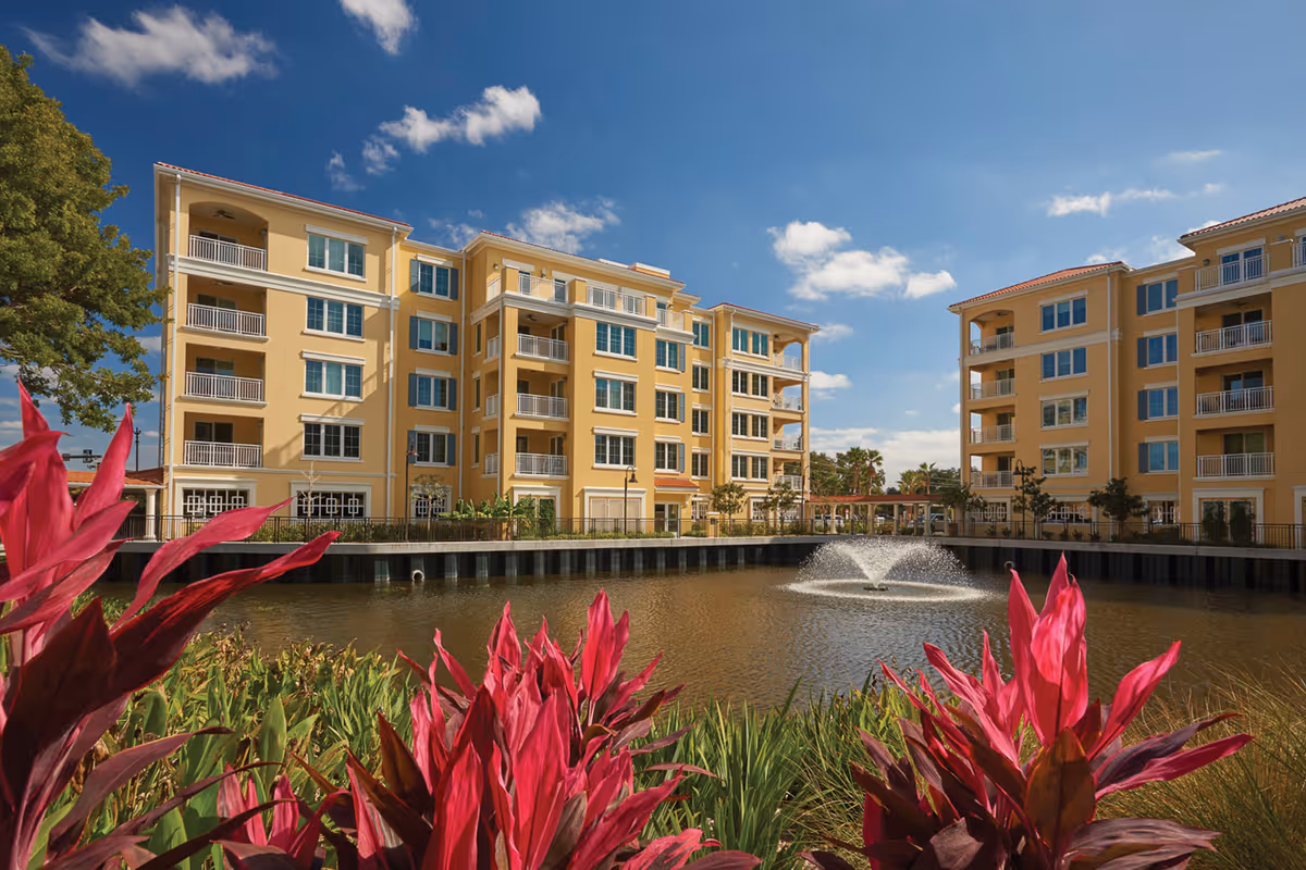 View of a senior living facility named Village On The Isle featuring multiple yellow multi-story buildings surrounding a pond with a water fountain, bright pink flowers and green plants in the foreground, and a clear blue sky with some clouds.