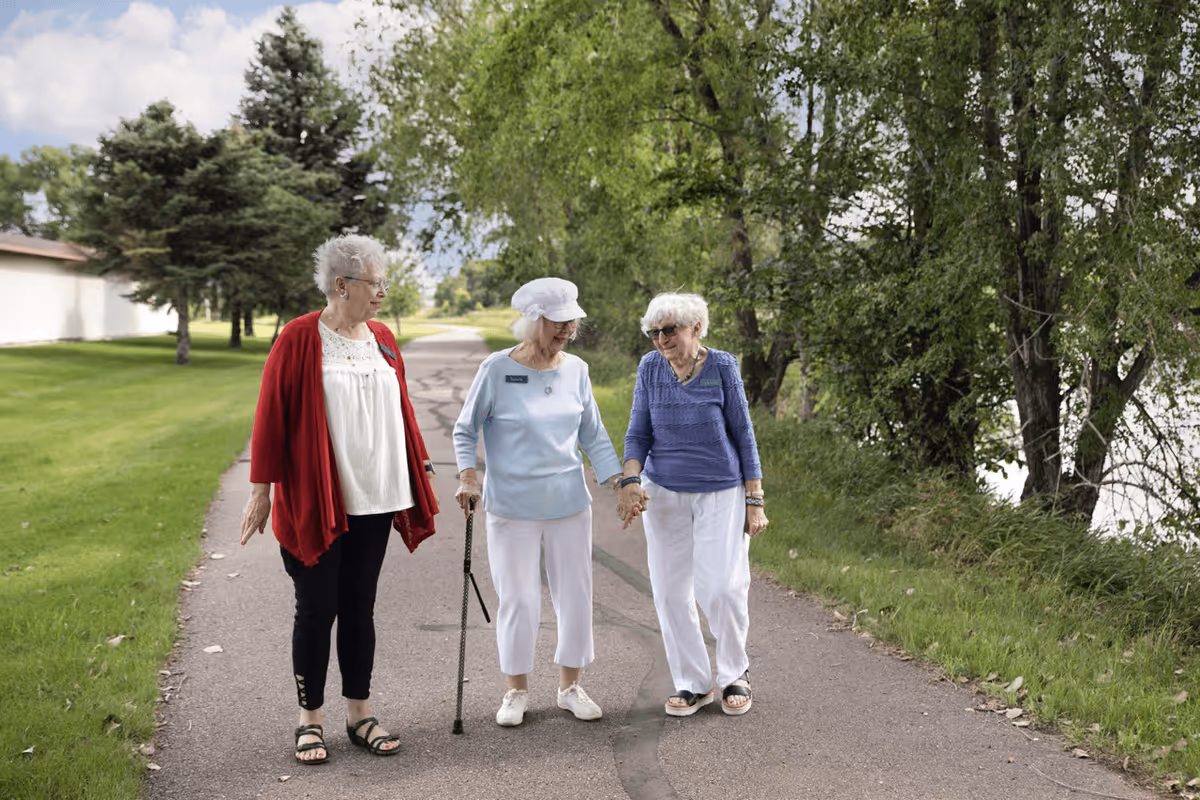 Three elderly women walking together on a paved path outdoors, surrounded by green grass and trees. One woman is wearing a red cardigan, another in a light blue top with a white hat and using a cane, and the third in a blue sweater holding hands with the woman using the cane.